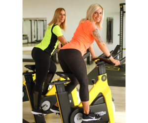 Fitnessforum Prenzlau – Zwei Frauen trainieren auf Spinningrädern in einem hellen Fitnessstudio und lächeln in die Kamera.